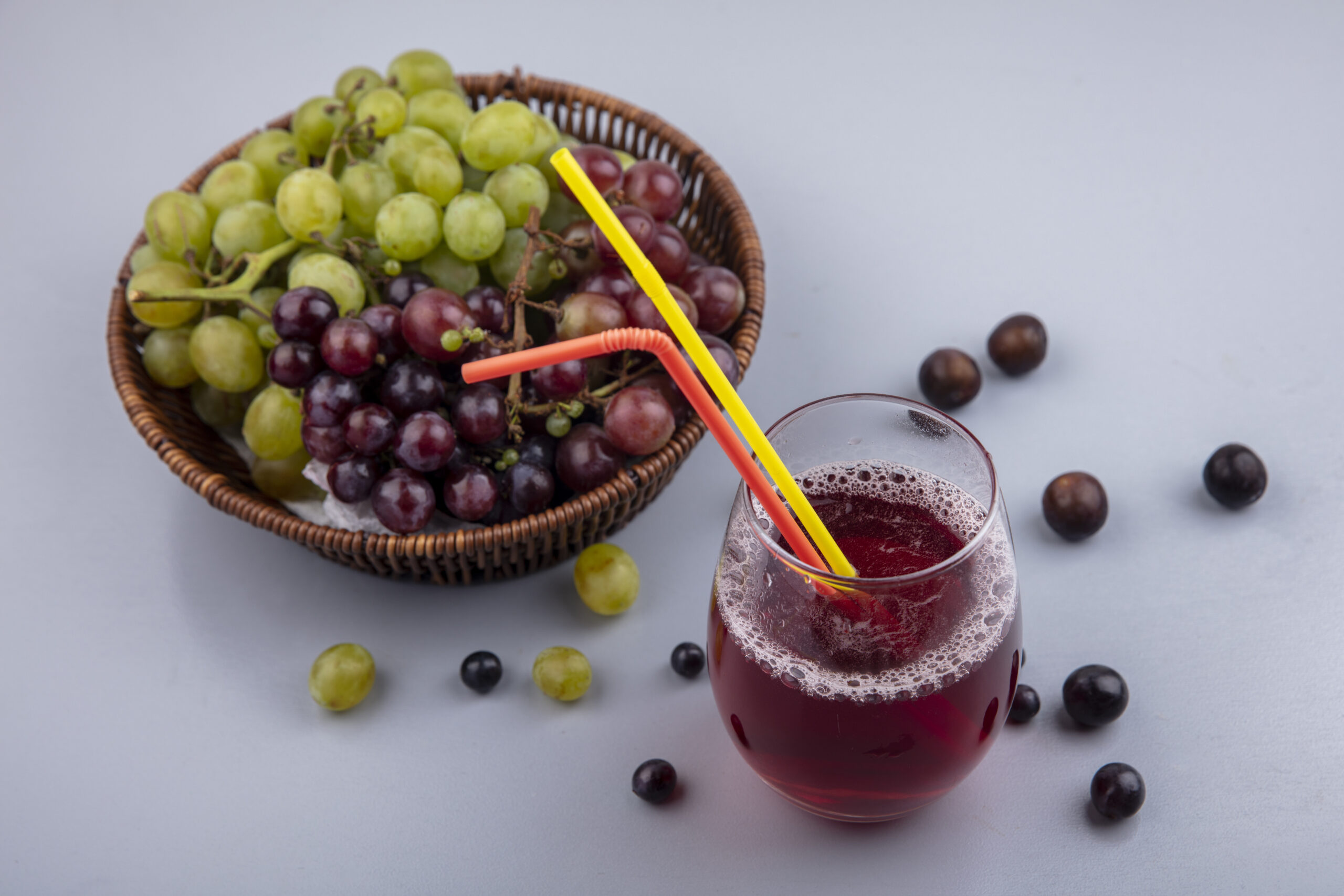 side view of black grape juice and drinking tubes in glass with grapes in basket and on gray background